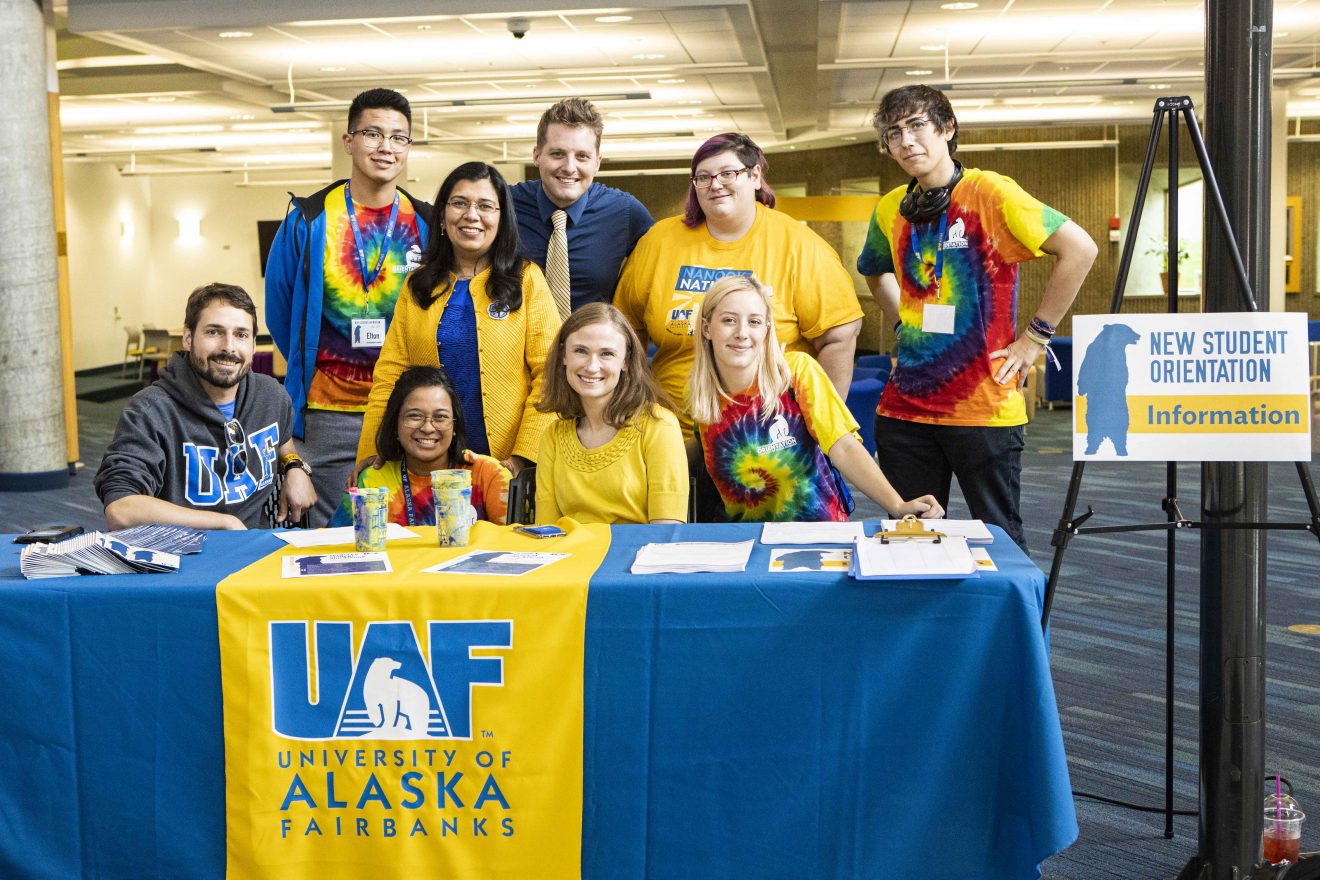 Provost Anupma Prakash stands with eight members of the New Student Orientation team behind an information desk in Wood Center.
