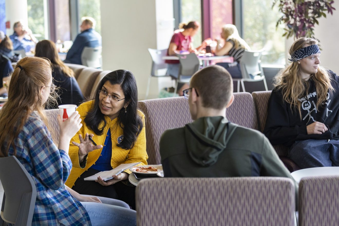 Anupma Prakash talks with two students over lunch at Dine 49. A third student sits off to the side, looking the other way. More people sit at other tables in the background.