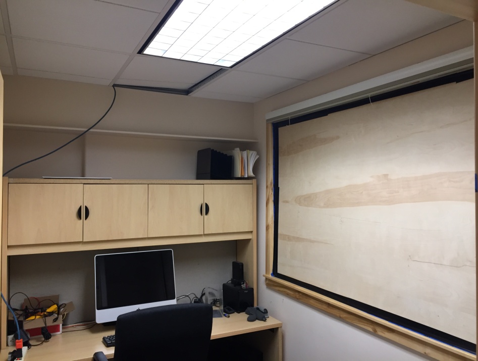 Picture of office with desk, chair, computer and cabinets. The long window to the side is complete blocked with a sheet of plywood. A panel of LED lights is overhead.