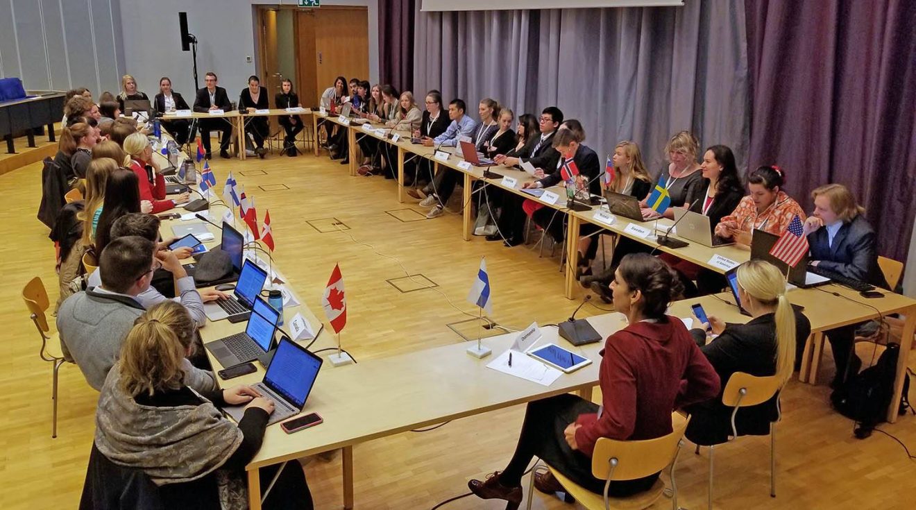 A few dozen students sit at tables in a rectangle to simulate an Arctic Council meeting. Flags or the various Arctic nations are in front of many of the students.