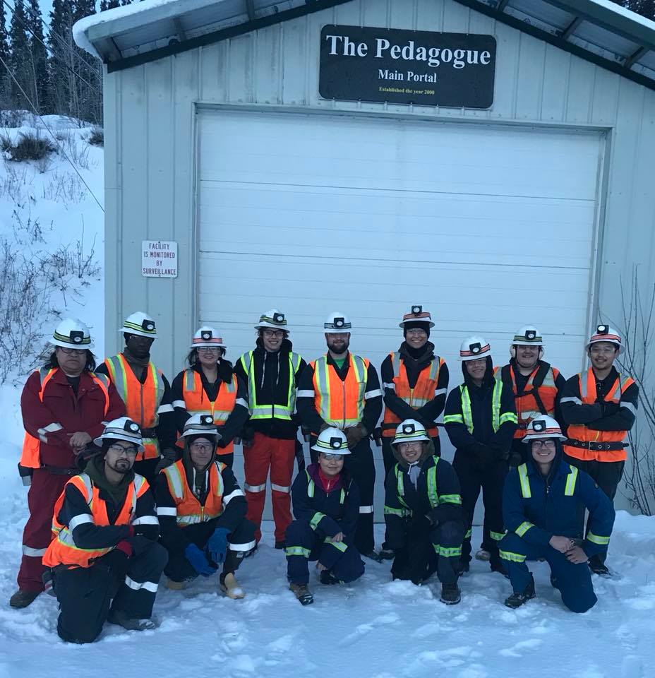 A group of people in hard hats and winter gear stand or kneel in the snow outside a structure that looks like a small garage or storage building.