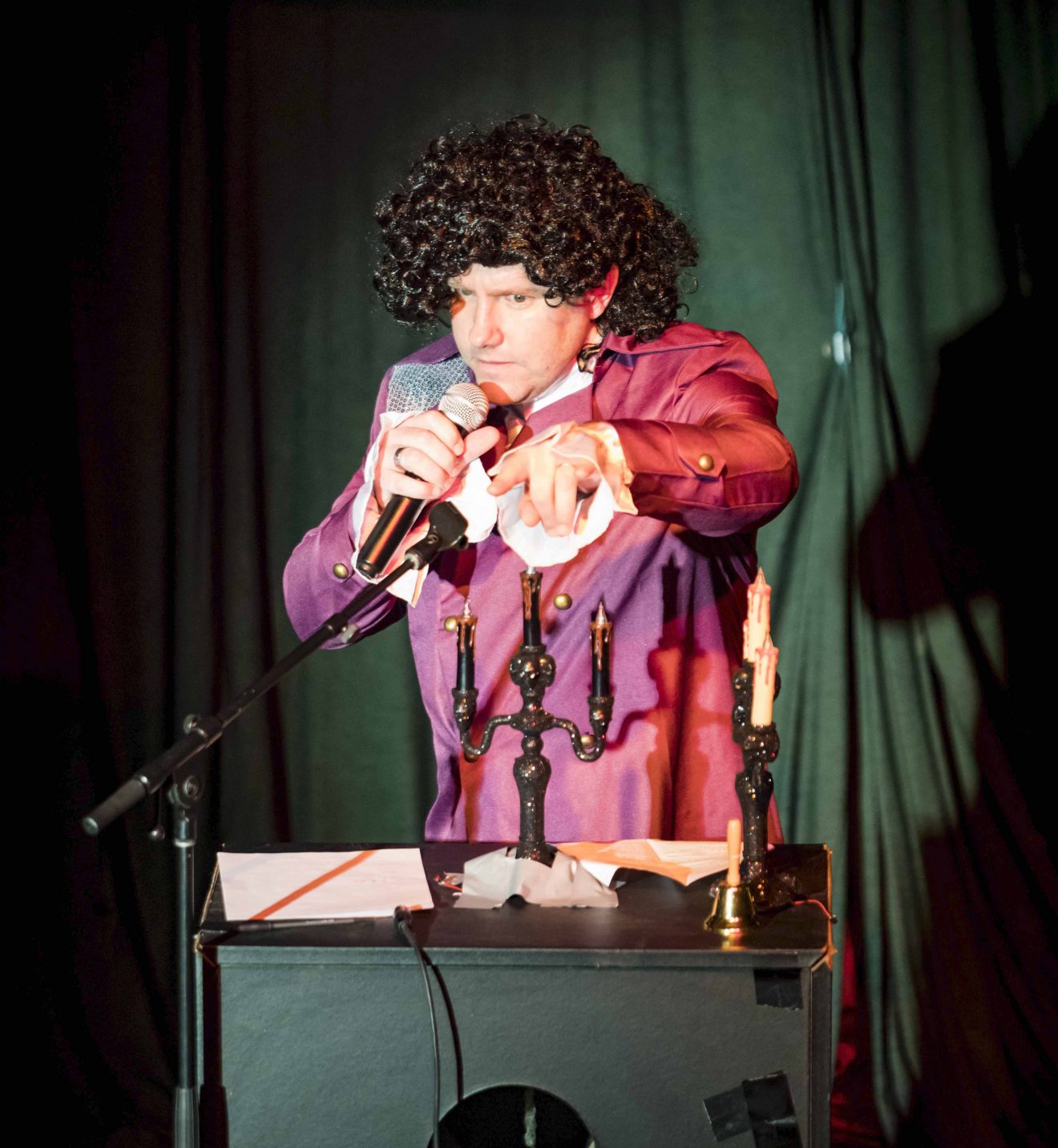 A man dressed as Prince speaks into a microphone. He is wearing a purple costume with long lace sleeves and a curly black wig.