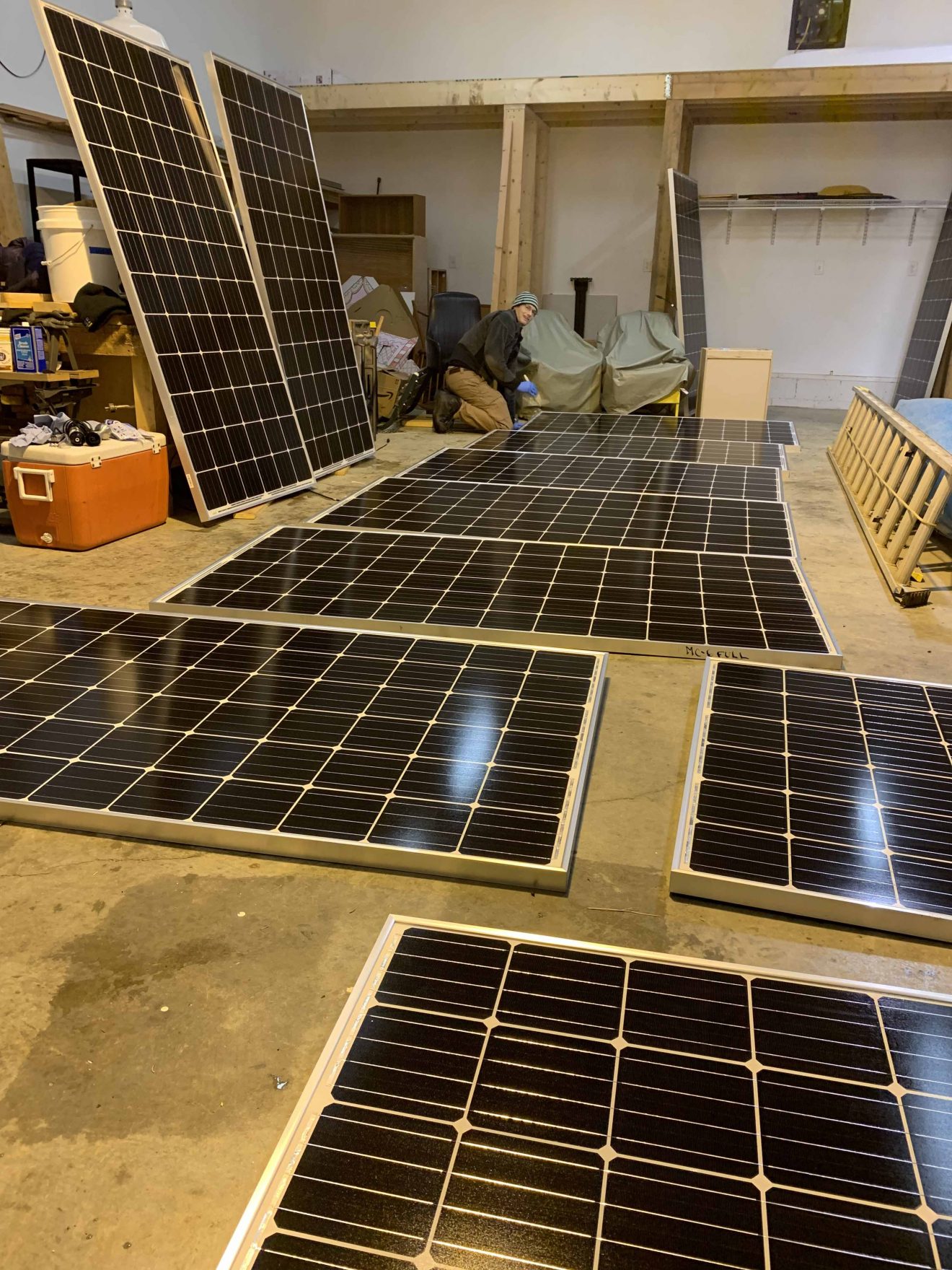 A room filled with solar panels on the floor and leaning against a bench. A person kneels in the background.