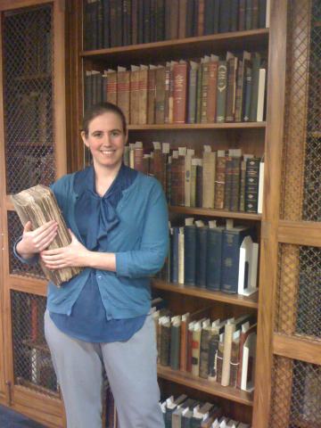Rachel Cohen stands in front on a tall bookshelf of old books and holds a large folio of old papers.
