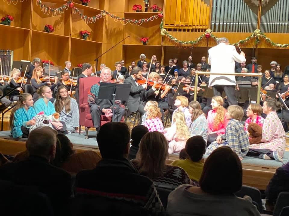 Children sit on the floor in front of a man in an armchair. An orchestra and choir are behind them.