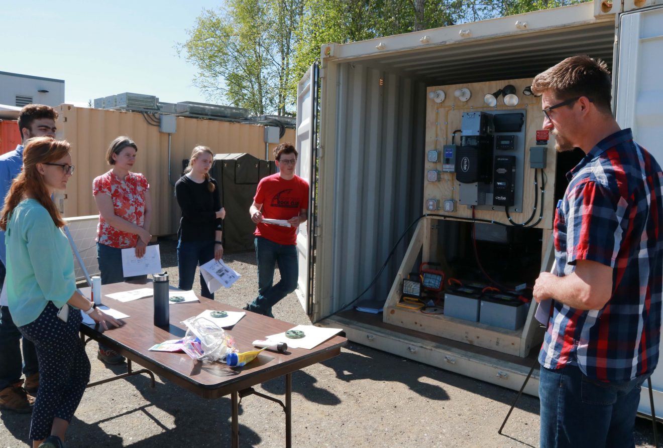 Six people stand around a folding table with papers, water bottles and miscellaneous things on it. An open freight container holds a display with a variety of batteries, lights and fuse boxes.