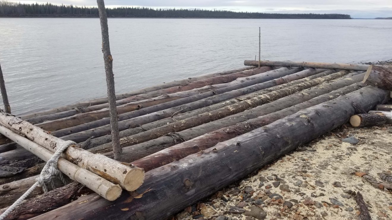 Raft of logs sits on a sandy gravel bar along a wide river.