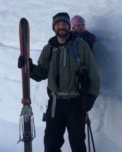 Photo of man holding a pair of skis. He is carrying a baby on his back.