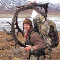 Larry Bartlett carrying caribou antlers and a backpack. He is on the tundra in fall. A river or lake is behind him.