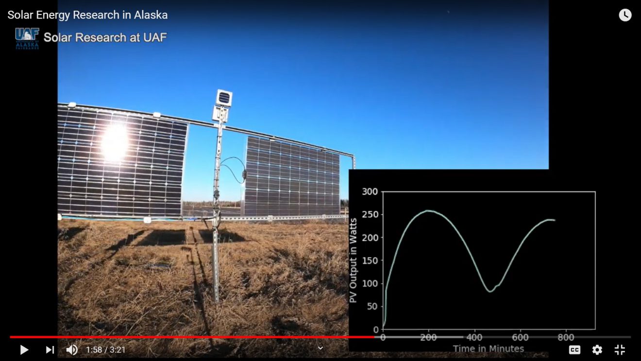 Screen shot of video still featuring solar panels and a computer screen with a graph on it.