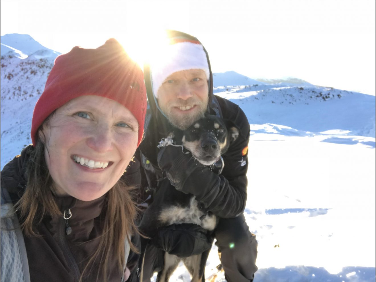 A woman and a man stand on a glacier with the sun directly behind them. They are in winter gear. The mean is holding a black and tan dog.