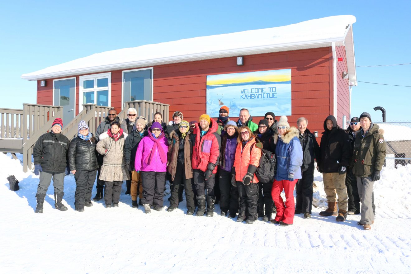 Large group of people bundled up in winter gear stand outside a small red house in the snow on a sunny day.