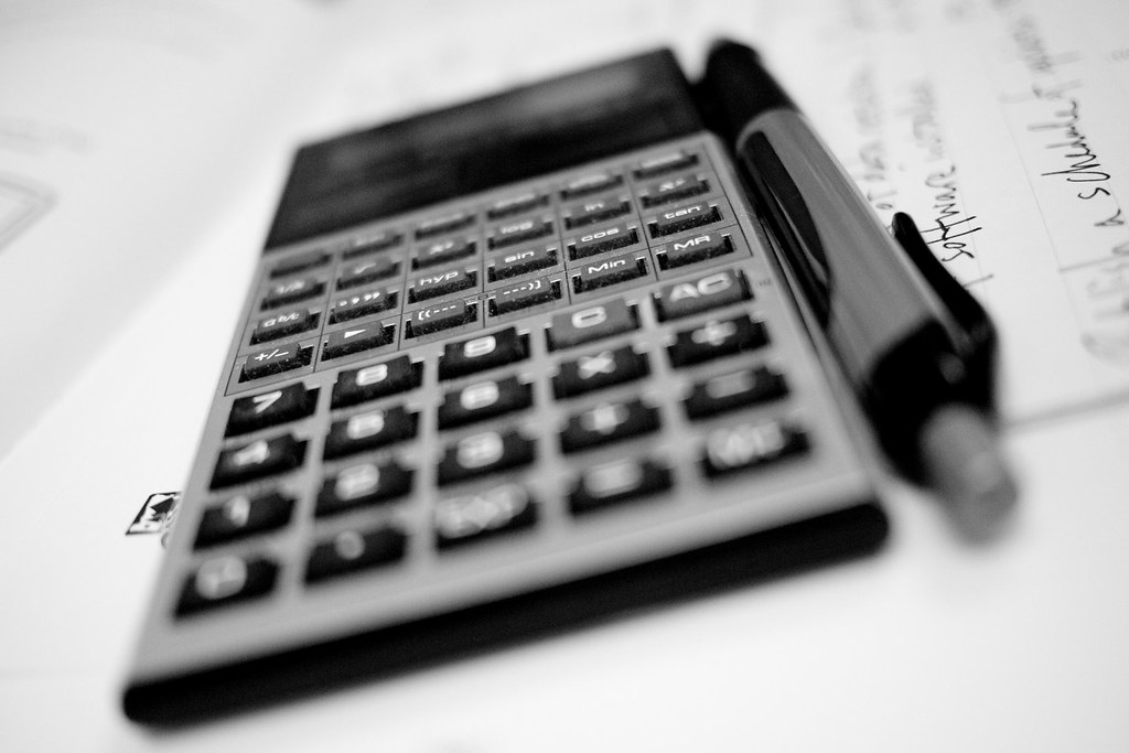 Close-up image of a calculator and pen lying on top of a piece of paper with writing on it. The writing is unclear.