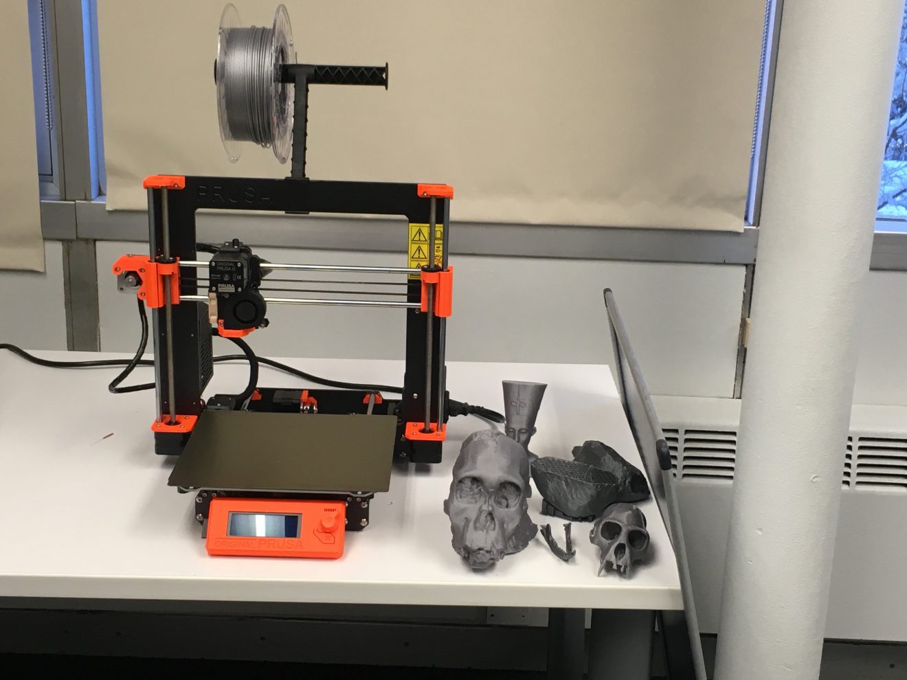Photo of a 3D printer. Next to it are model printouts of two skulls, a small Egyptian bust, what appears to be a jawbone and two other items that are not clearly discernible.