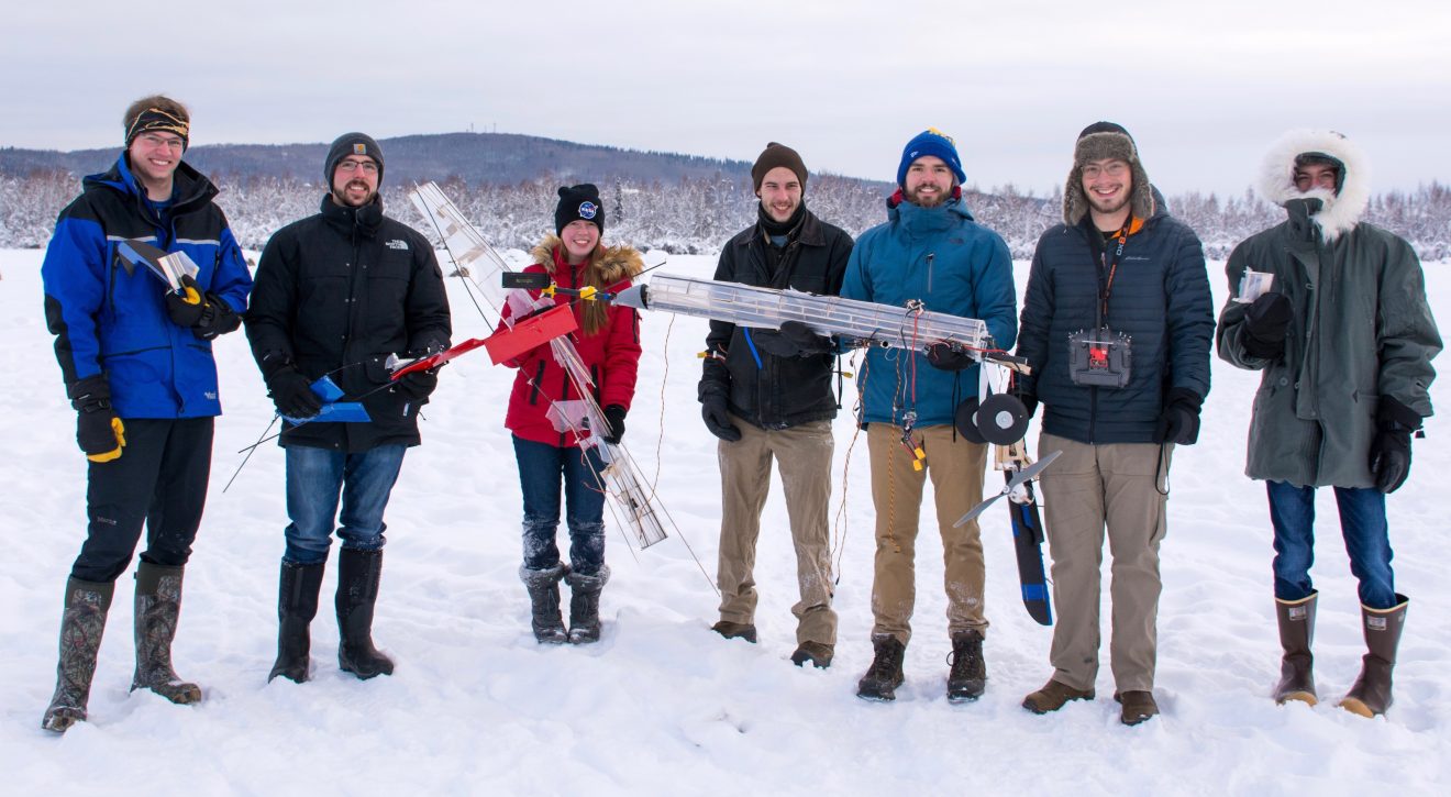 A group of people stand in the snow. Some of them are holding what appear to be small planes, probably drones. Others are holding small items that may be controllers or computer components.