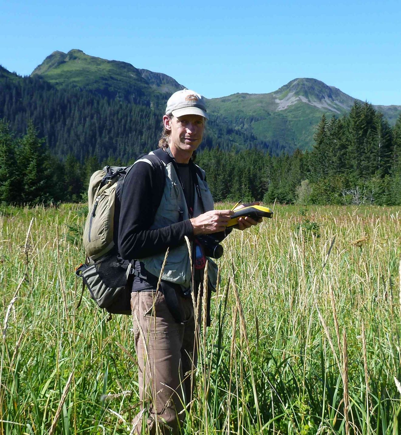 Photo of man standing in a field of grass with mountains behind him. He is carrying a backpack and holds a notebook and pencil in his hands.