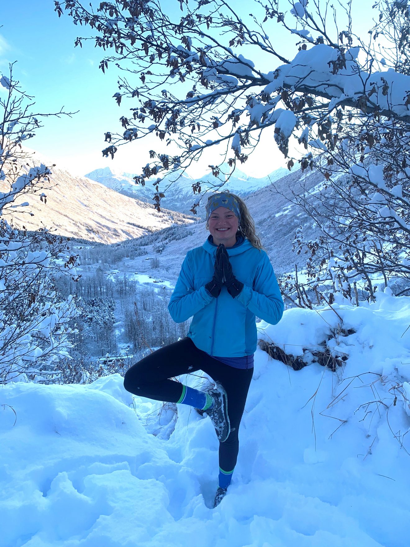 A young woman holds a yoga pose while standing in the snow. She is in winter clothing. A valley framed by mountains stretches behind her.