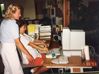 A woman standing behind a woman who is sitting at a large computer.