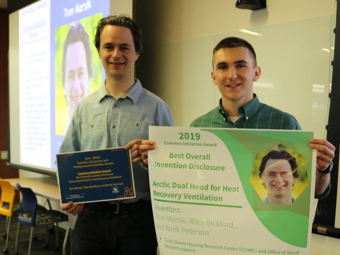 Two man stand holding an award certificate and a poster. They are in a classroom.