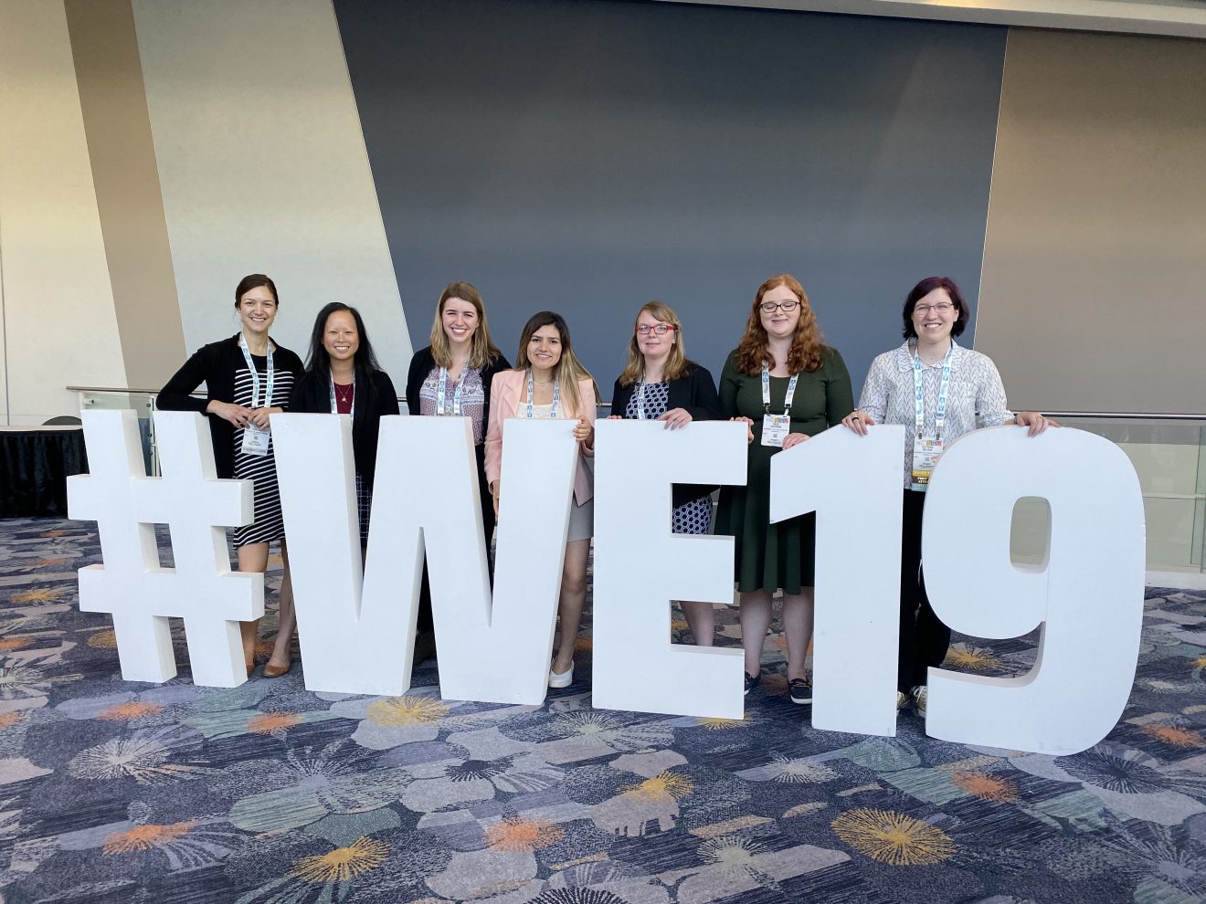 Seven women stand behind a large sign that reads "#WE19"