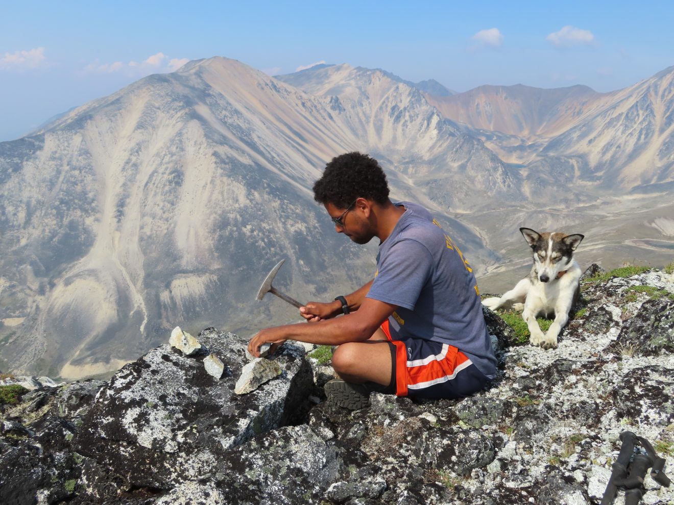 A man sits on a rocky ridge with a small hammer for a rock specimen. He is in the mountains. A dog sits behind him.