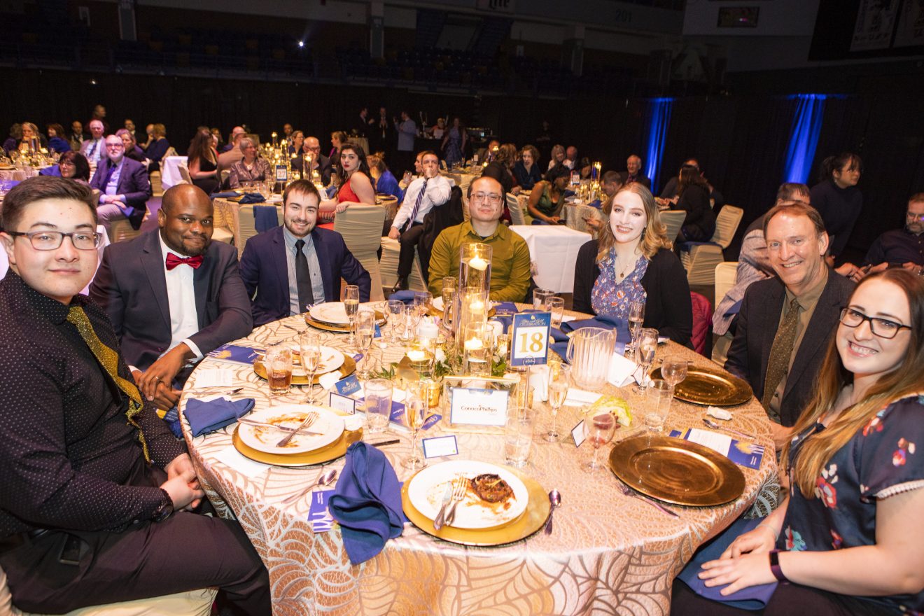 Seven people sit around a table in a room filled with other people. Everyone is dressed up. The tables are set with blue and gold decorations.