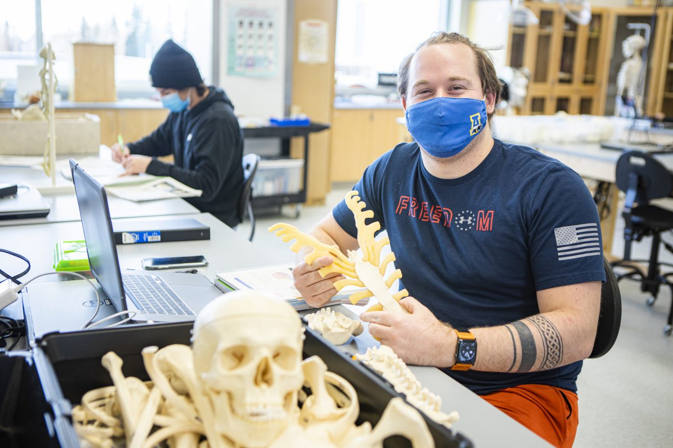 A man wearing a mask sits at a lab table holding what appears to be part of a skeleton, probably made of plastic. Another student, also wearing a mask, sits some distance away.