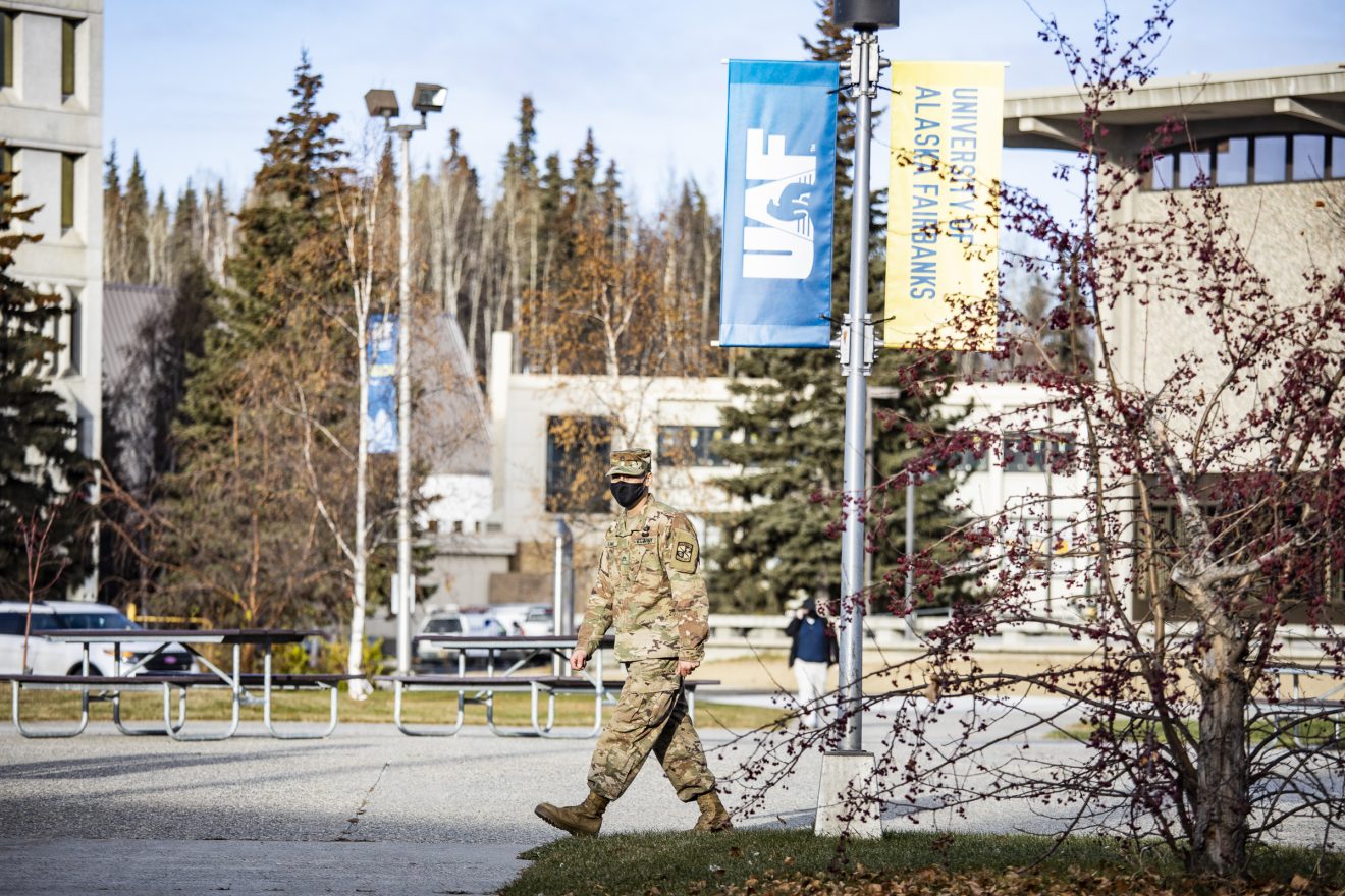 Man in an Army uniform and wearing a mask walks across campus. Some buildings are behind him, and most of the leaves have fallen from the trees.