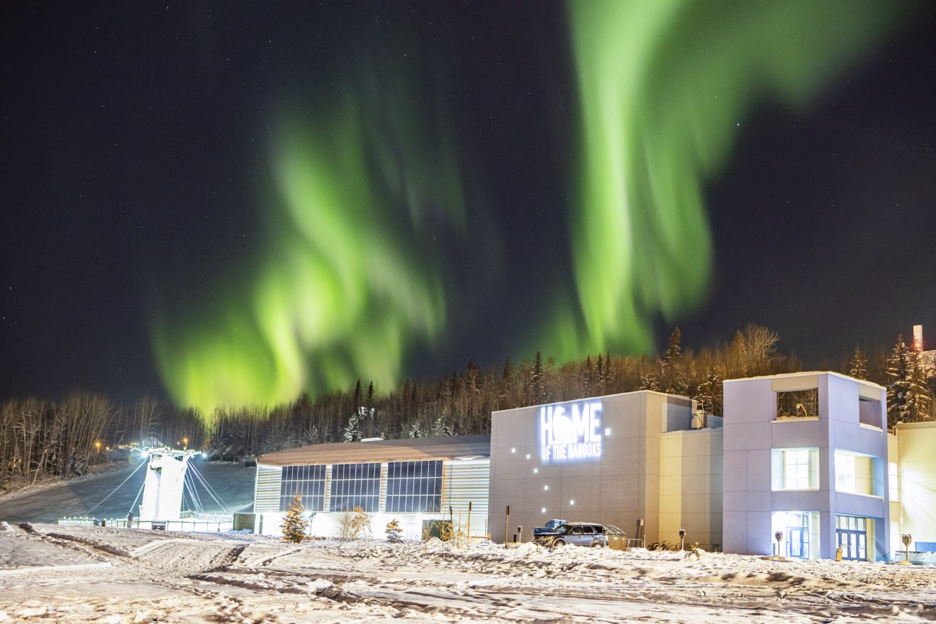 Night-time shot of a green aurora over a brightly lit building. "Home of the Nanooks" logo is lit on one side. It is winter.