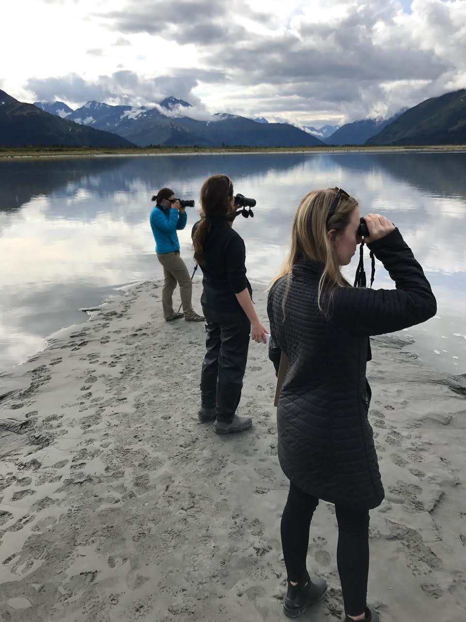three people stand on a beach looking out at the water with binoculars