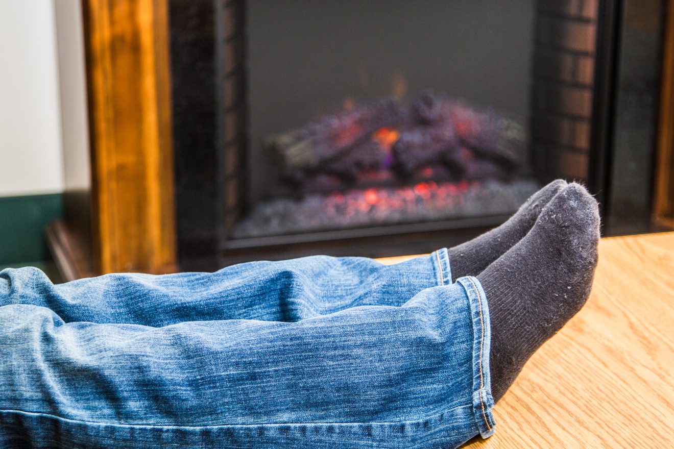 Pair of legs wearing jeans and warm socks are stretched out on a footstool in front of an electric fire.