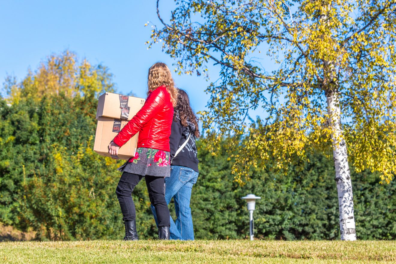 Two students walk across the lawn on a sunny fall day. One is carrying two cardboard boxes.