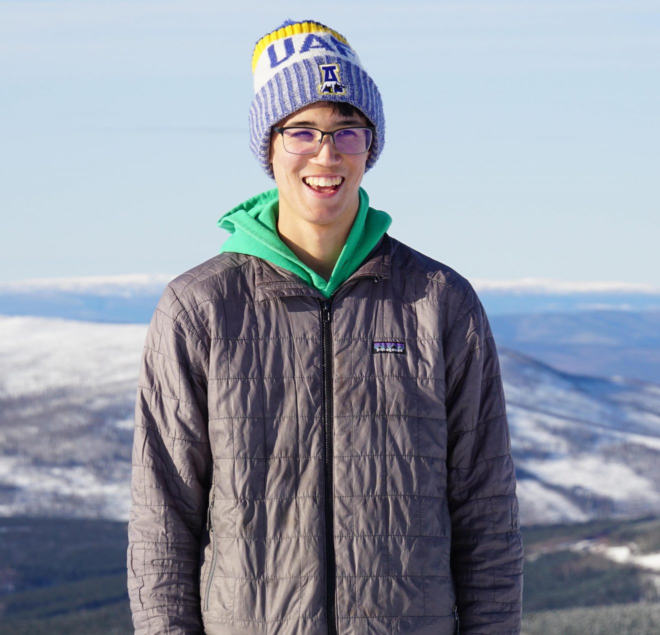 Photo of a young man in light winter clothing standing atop a hill. Snow-covered hills and mountains stretch behind him.