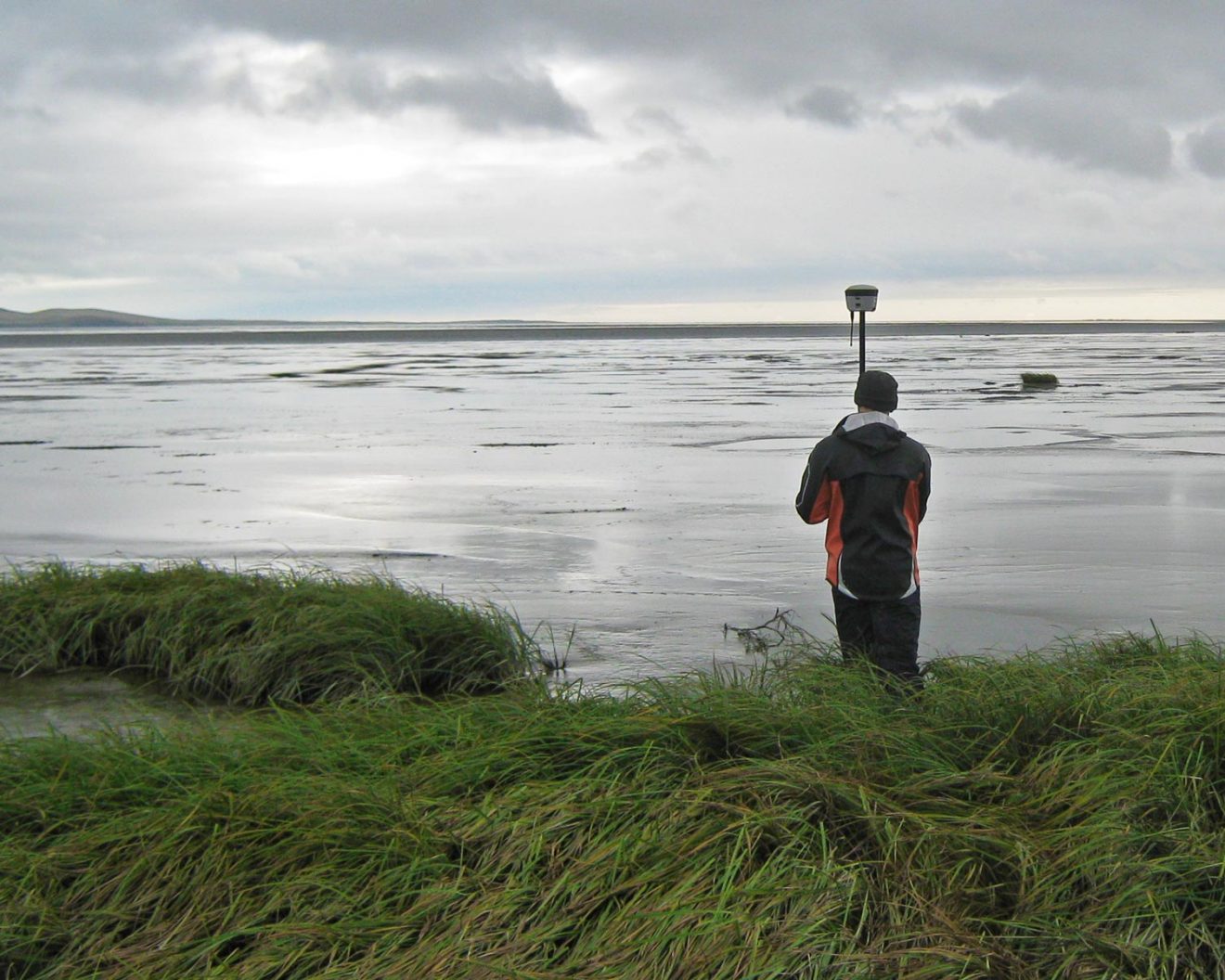 A man stands in long grass by the edge of the ocean. He is hold a tall pole with a small box attached to the top, possibly a camera or a measurement device.