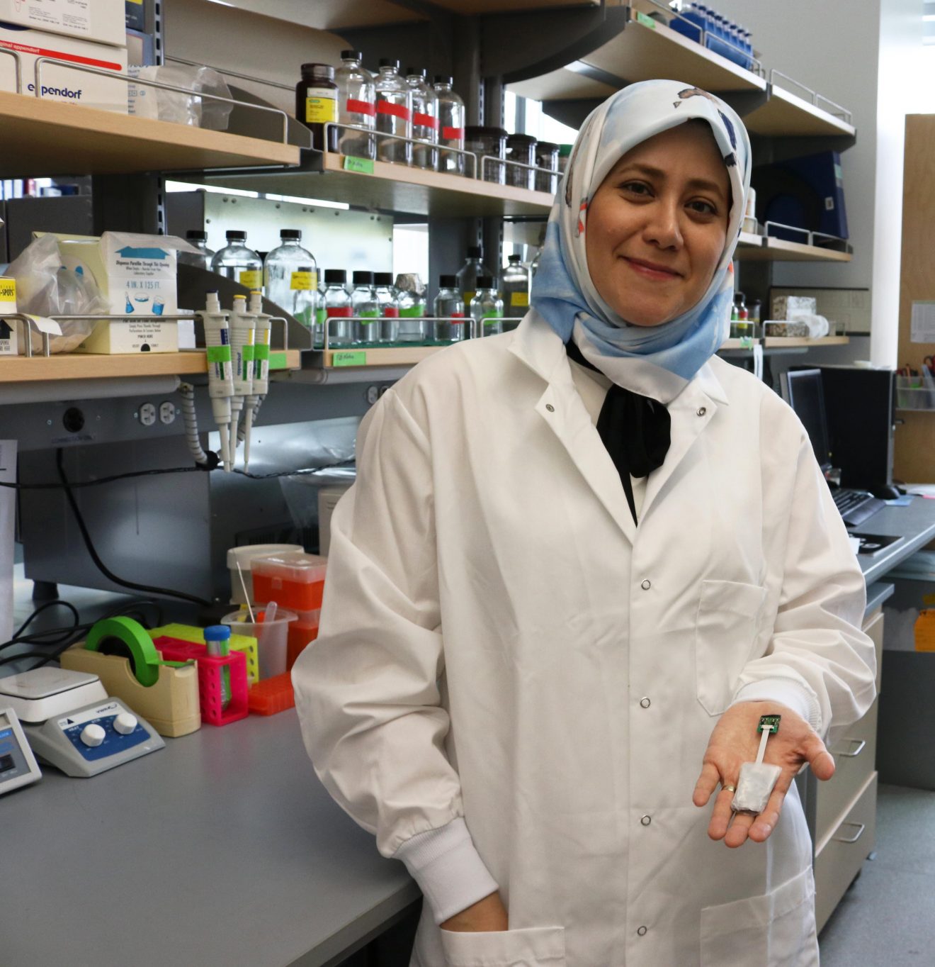 Photo of a woman in a lab coat, standing in a lab, holding a small device in her hand.