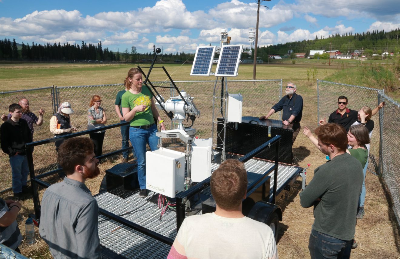 A group of people stand outside around some small solar panels and equipment presumably related to the panels.