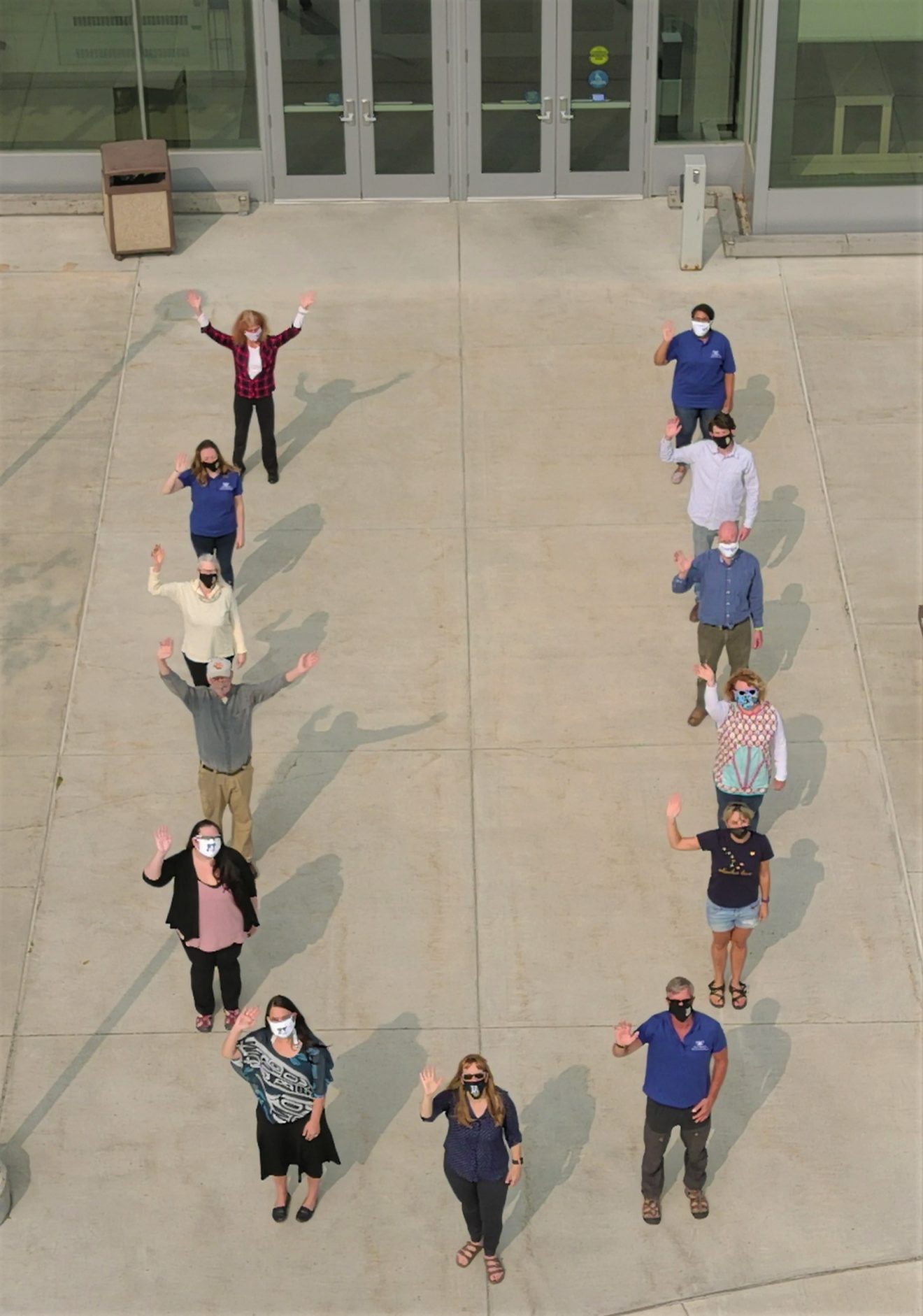 13 people stand outside to form a U as seen from a drone flying above. They are wearing masks (COVID-19).