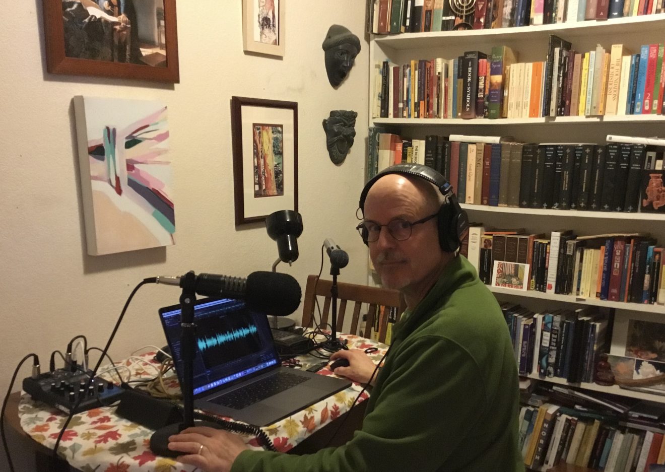 A man with headphones sits in front of a microphone and a laptop in a room filled with artwork and books, presumably a makeshift studio.
