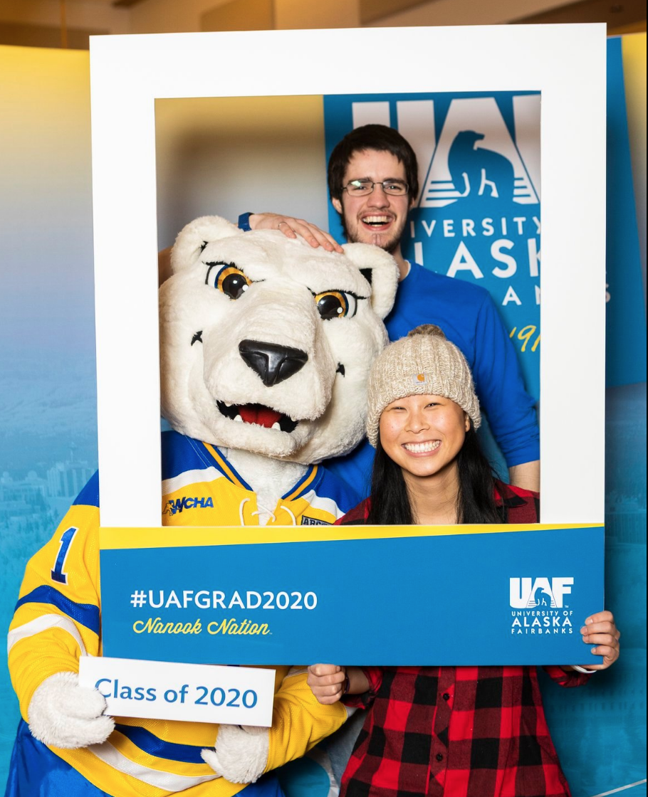 Two people plus the Nanook mascot pose behind a #UAFGrad2020 frame.