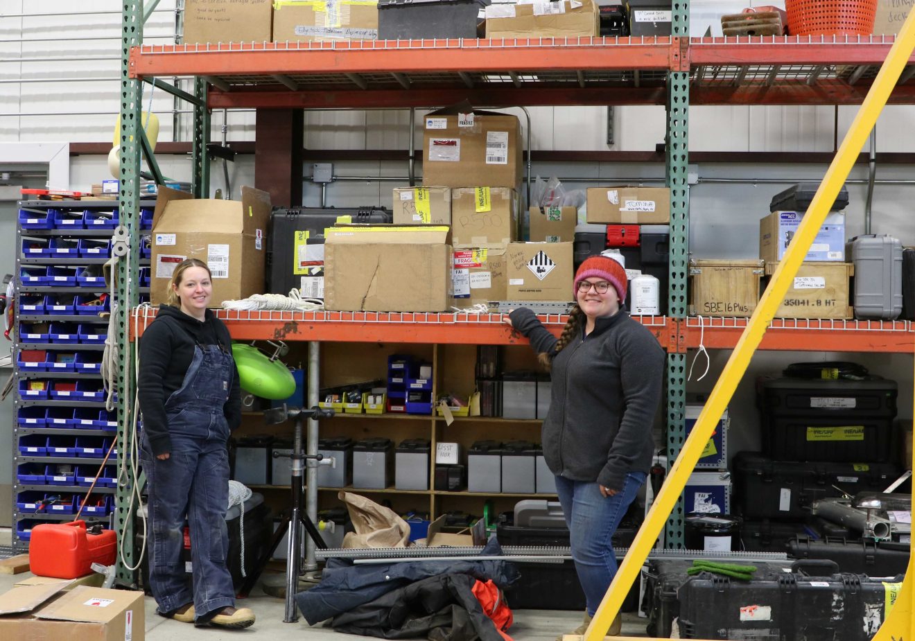 Two women stand in a warehouse in front of many shelves filled with boxes. Gear and gear boxes are piled around them.