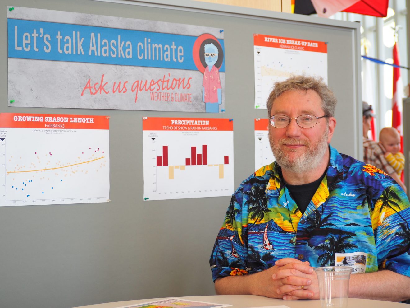 Photo of Rick Thoman sitting in front of a board with a sign inviting people to ask questions about Alaska climate.