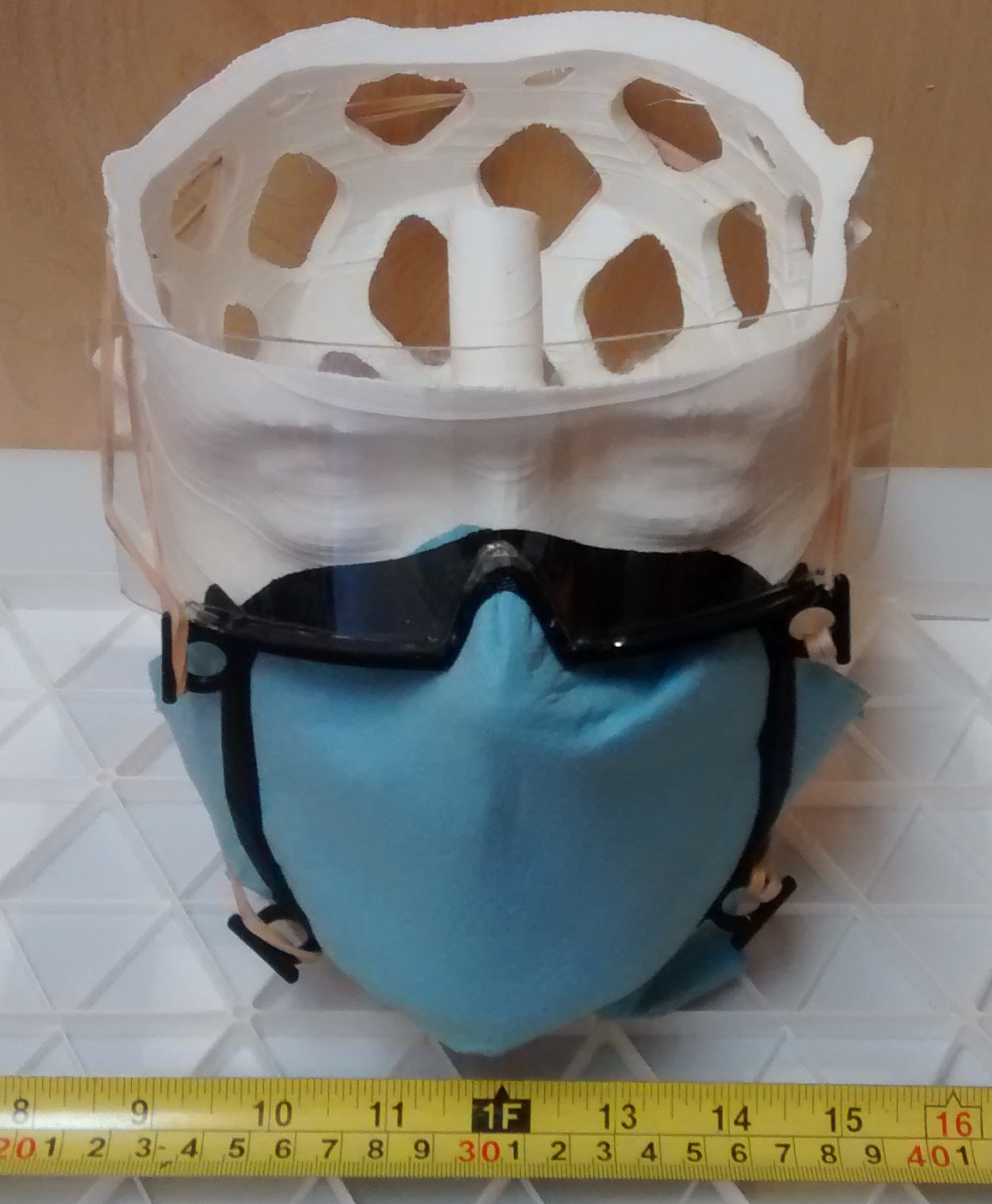 Sample mask wrapped around a skull-shaped frame, topped by an eye-protection shield. A tape measure shows the width is 6-7 inches wide.