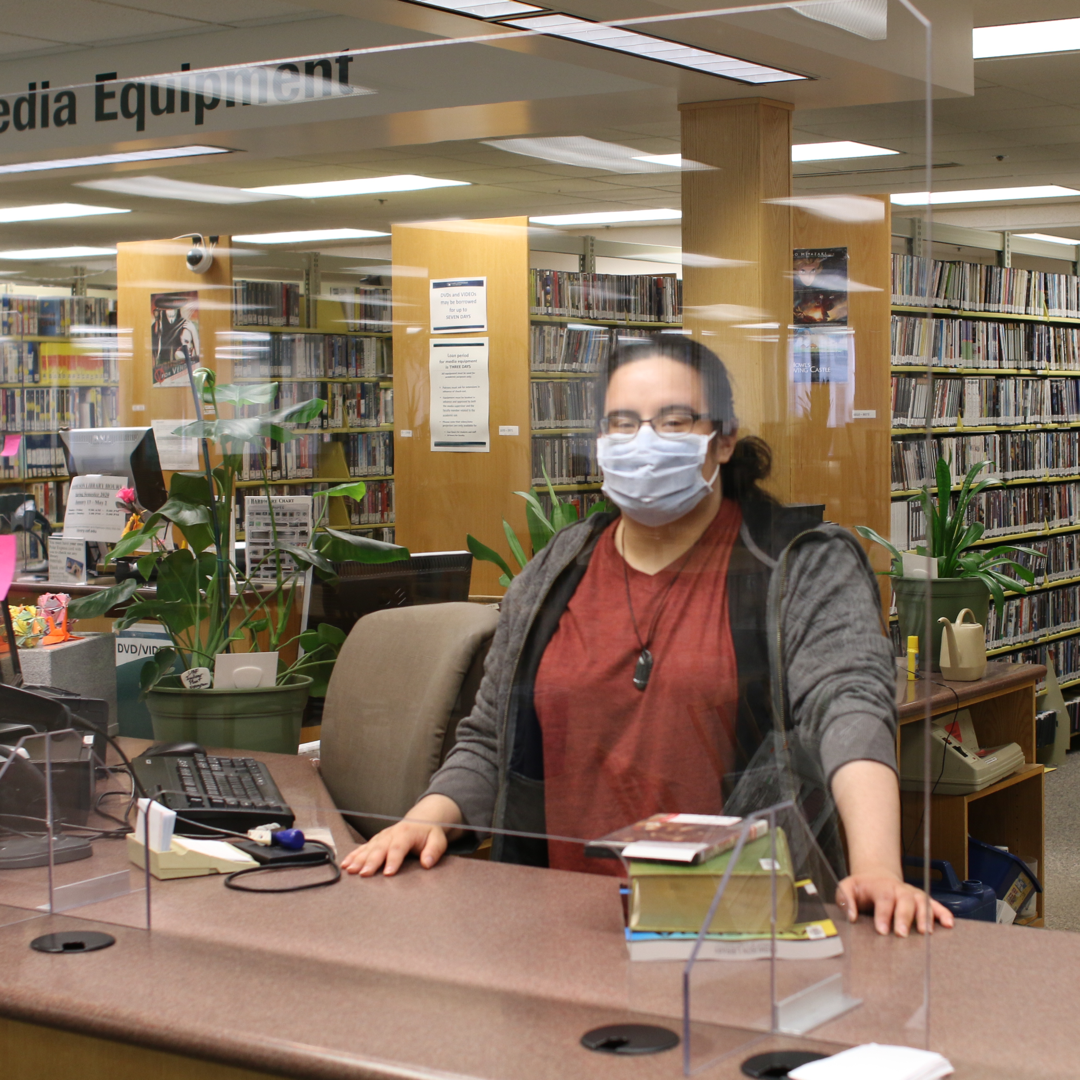 A student wearing a mask stands behind a desk. Behind her are many bookshelves filled with books.