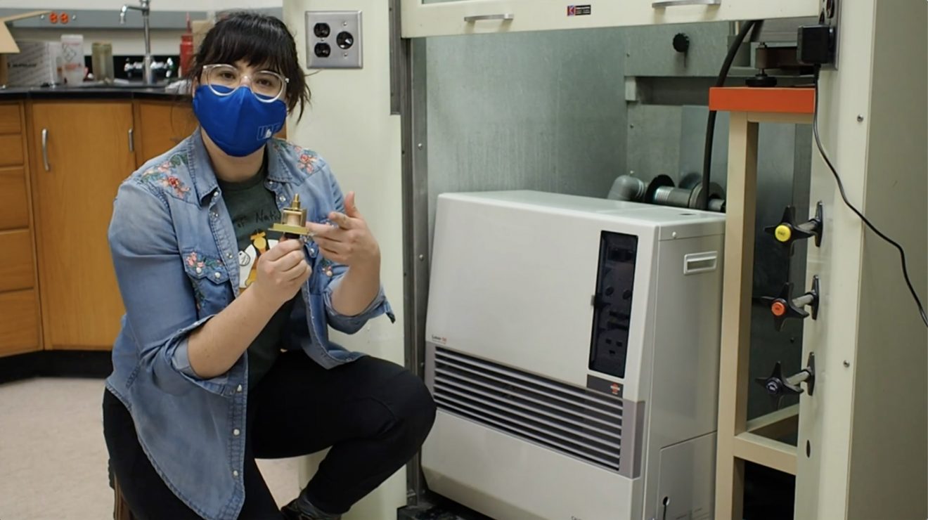Woman wearing a mask kneels by a home heating unit. She is holding a small device in her hand.