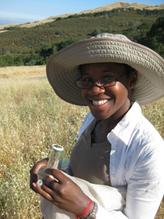 A woman wearing a sun hat stands in a field. She is holding a vial or small container.