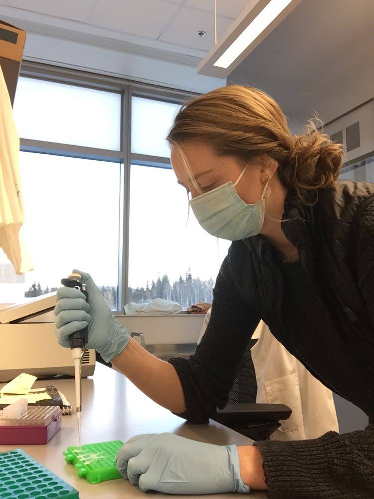 A woman wearing a mask and gloves uses a pipette over a small tray.