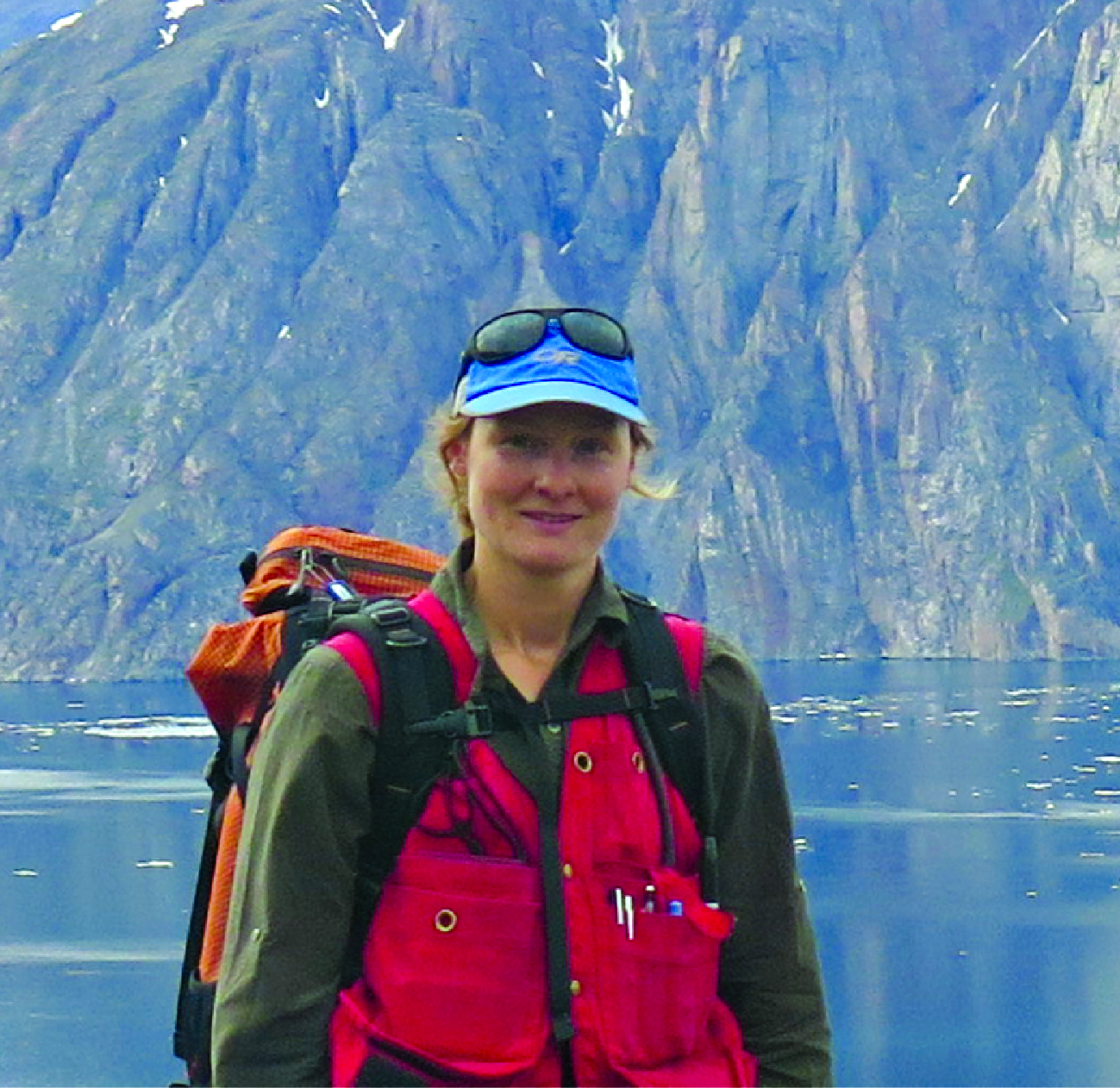Photo of woman outside. She is wearing a backpack and stands in front of a body of water and a mountain.