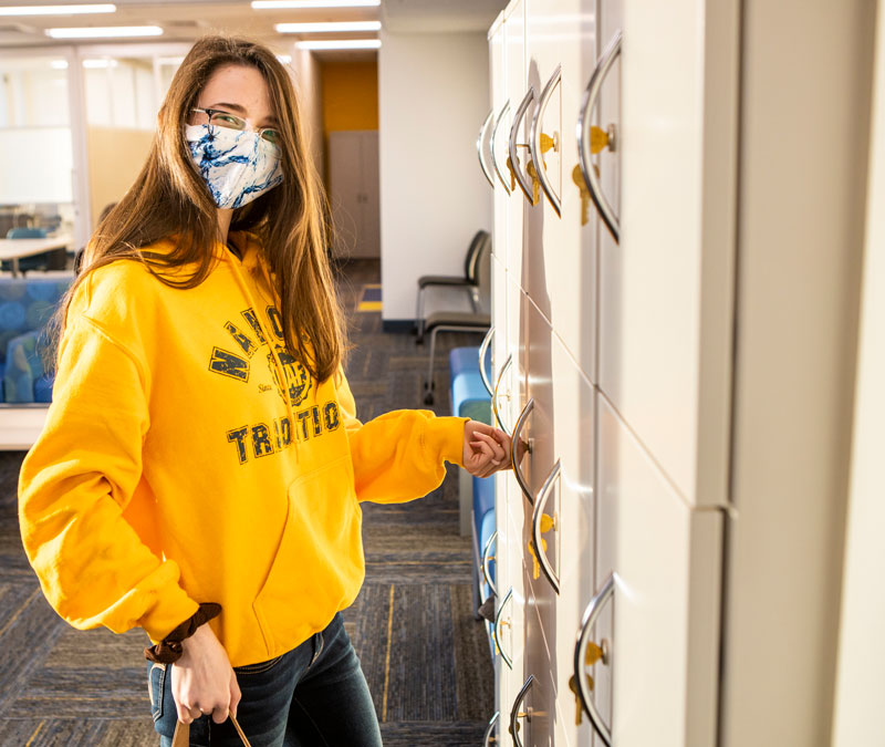 Young lady in yellow sweatshirt and mask turning a key in a locker.
