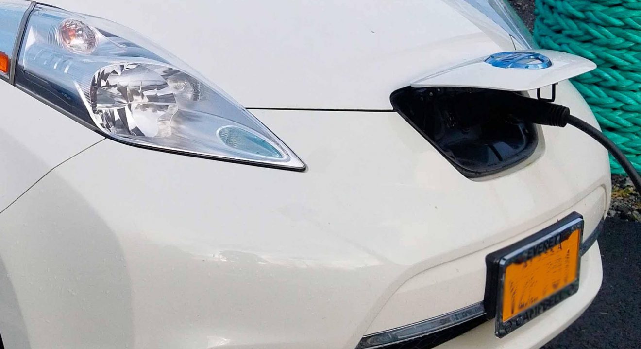 Closeup of the hood of an electric car with a electrical cord plugged into it.
