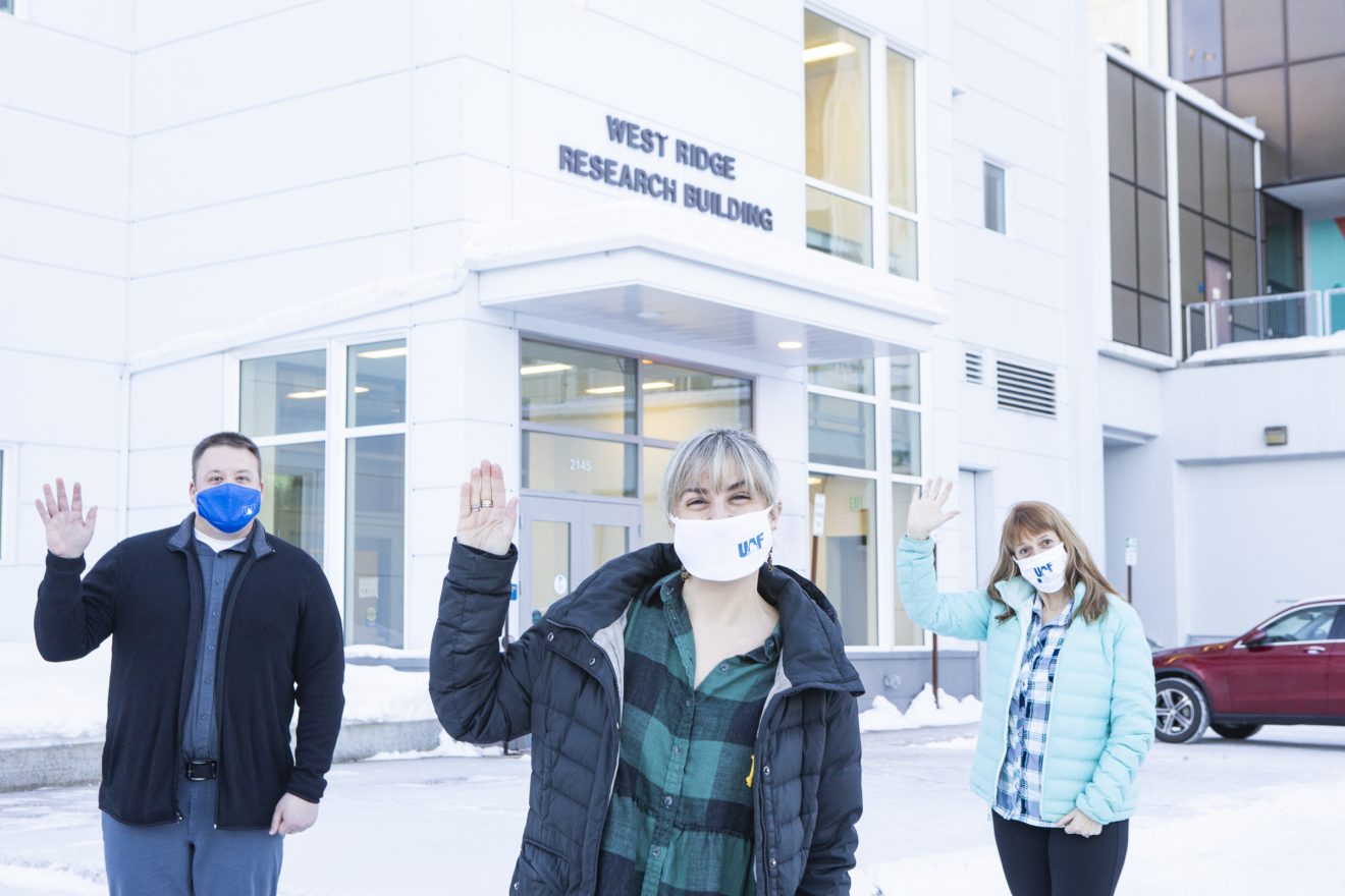 Three people wearing masks and physically distanced stand outside a building in winter. They are waving at the camera.